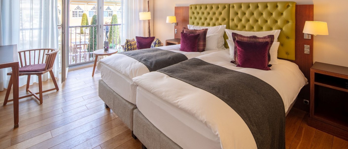 Ein helles Doppelzimmer mit zwei Betten, lila Kissen, Holzboden und einem Balkon mit Blick auf einen Innenhof., © Waldhotel Stuttgart GmbH