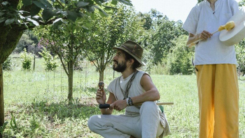 Zwei Personen im Garten, eine kniet mit einem Instrument, die andere steht mit einem Hut. Sie sind von Bäumen umgeben und es herrscht eine entspannte Atmosphäre., © Theaterhaus Stuttgart e.V.