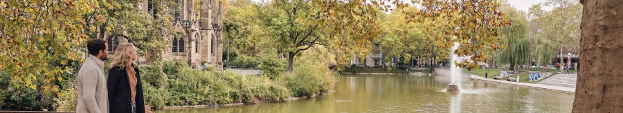 Ein Paar spaziert am Feuersee entlang, im Hintergrund die Johanneskirche und herbstliche Bäume. Ein Brunnen sprudelt im See., © SMG Christoph Düpper Ein Paar spaziert am Feuersee entlang, im Hintergrund die Johanneskirche und herbstliche Bäume. Ein Brunnen sprudelt im See., © SMG Christoph Düpper