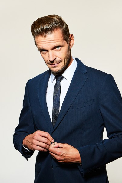 A man in a dark blue suit buttons up his jacket and looks confidently into the camera. The background is neutral., &copy; Theaterhaus Stuttgart e.V.
