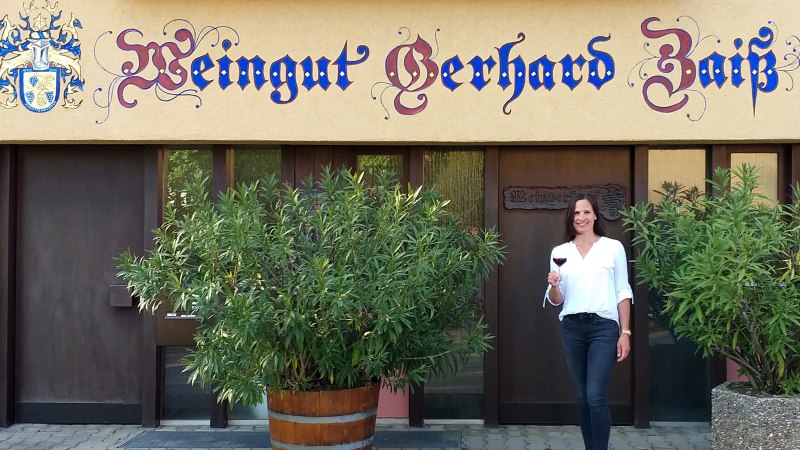 Eine Frau steht vor dem Weingut Gerhard Zaiß, hält ein Glas Rotwein und lächelt. Das Gebäude hat eine dekorative Beschriftung und Pflanzen im Vordergrund., © Simone Mack Eine Frau steht vor dem Weingut Gerhard Zaiß, hält ein Glas Rotwein und lächelt. Das Gebäude hat eine dekorative Beschriftung und Pflanzen im Vordergrund., © Simone Mack