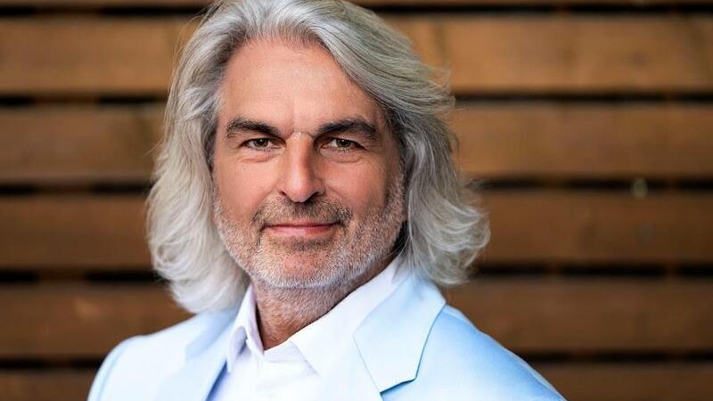 Ein Mann mit grauem Haar und Bart trägt einen weißen Anzug. Er steht vor einem Holzhintergrund und lächelt leicht., © Rosenau Kultur e.V. Ein Mann mit grauem Haar und Bart trägt einen weißen Anzug. Er steht vor einem Holzhintergrund und lächelt leicht., © Rosenau Kultur e.V.