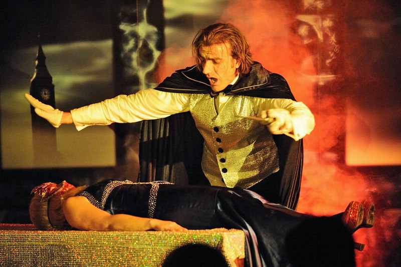 Actor in Victorian costume with cloak on stage, one person lying on a table, Big Ben in the background., © Gruseldinner GmbH & Co. KG Actor in Victorian costume with cloak on stage, one person lying on a table, Big Ben in the background., © Gruseldinner GmbH & Co. KG