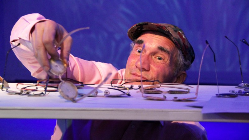 A man in a cap smilingly reaches for a pair of glasses on a table covered with several pairs of glasses. The background is illuminated in blue., &copy; Katja Ritter