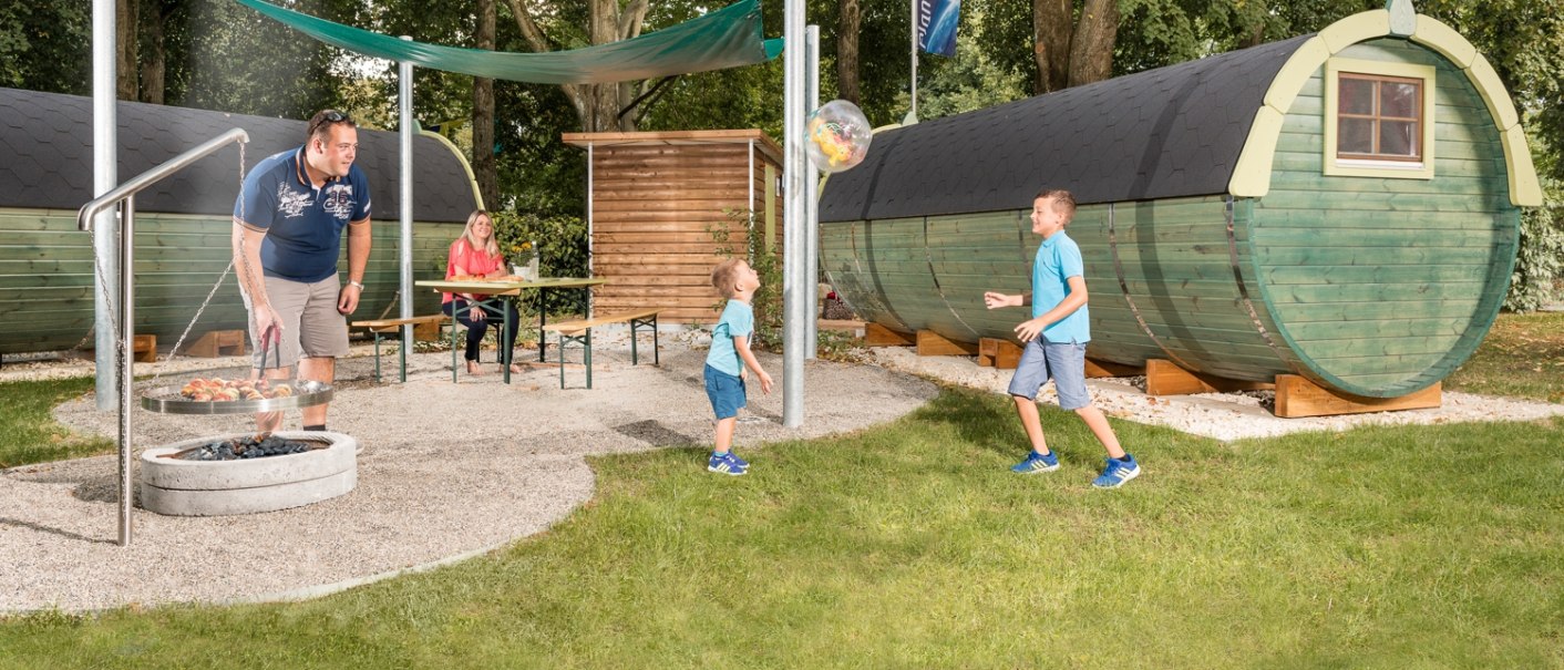Familie genießt Grillplatz mit Fasshütten im Hintergrund. Ein Mann grillt, während Kinder spielen und eine Frau am Tisch sitzt., © Remsfässer