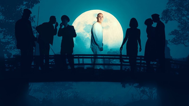 Ein Mann in einem Mantel und Hut sitzt mit ernstem Blick. Das Bild ist in Schwarz-Weiß gehalten., © tri-bühne e.V. Ein Mann in einem Mantel und Hut sitzt mit ernstem Blick. Das Bild ist in Schwarz-Weiß gehalten., © tri-bühne e.V.