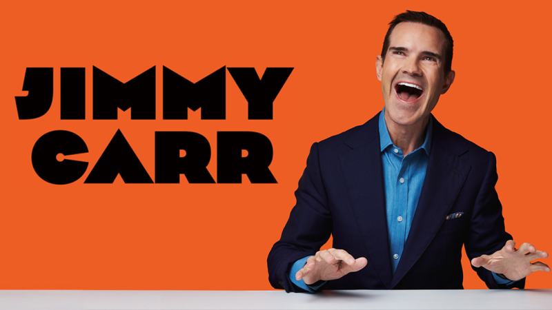 Ein Mann in einem blauen Hemd und dunklem Anzug lacht vor einem orangefarbenen Hintergrund mit dem Schriftzug 'Jimmy Carr'., &copy; links im Bild