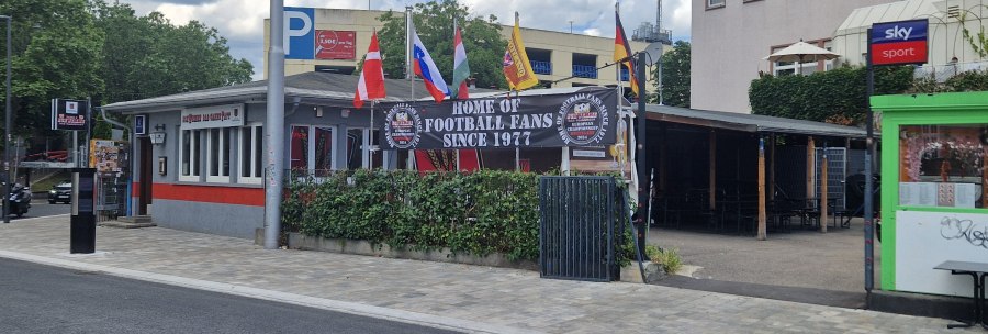 Bar mit Fahnen und Schild "Home of Football Fans Since 1977". Gebäude mit roten und grauen Akzenten, umgeben von Bäumen und einem Parkplatz., © Stuttgart-Marketing GmbH