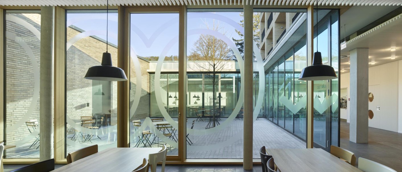 A modern restaurant with wooden tables and chairs; large windows offer a view of a courtyard with trees and seating areas., &copy; TOMAS