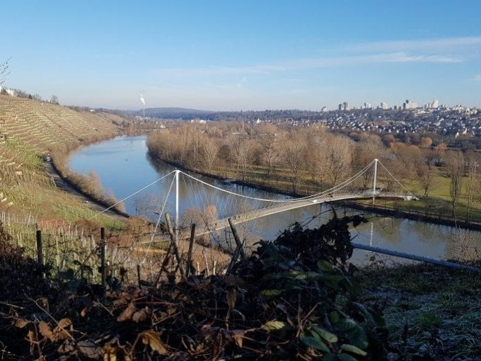 Hängebrücke über einen Fluss, umgeben von Weinbergen und kahlen Bäumen, mit einer Stadt im Hintergrund unter klarem Himmel., © Cool-Tours StattReisen Hängebrücke über einen Fluss, umgeben von Weinbergen und kahlen Bäumen, mit einer Stadt im Hintergrund unter klarem Himmel., © Cool-Tours StattReisen