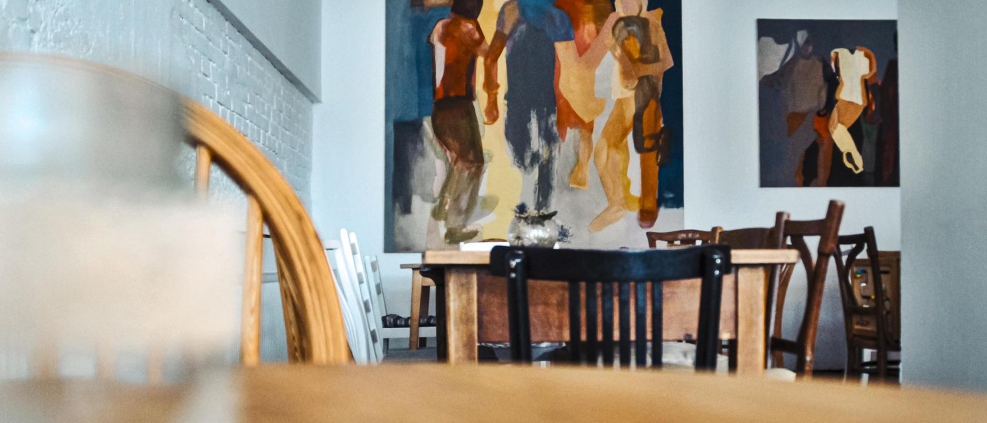 Ein gemütlicher Caféraum mit Holztischen und Stühlen, an der Wand hängen abstrakte Gemälde. Der Raum wirkt einladend und künstlerisch., © Raupe Immersatt e.V., Stuttgart