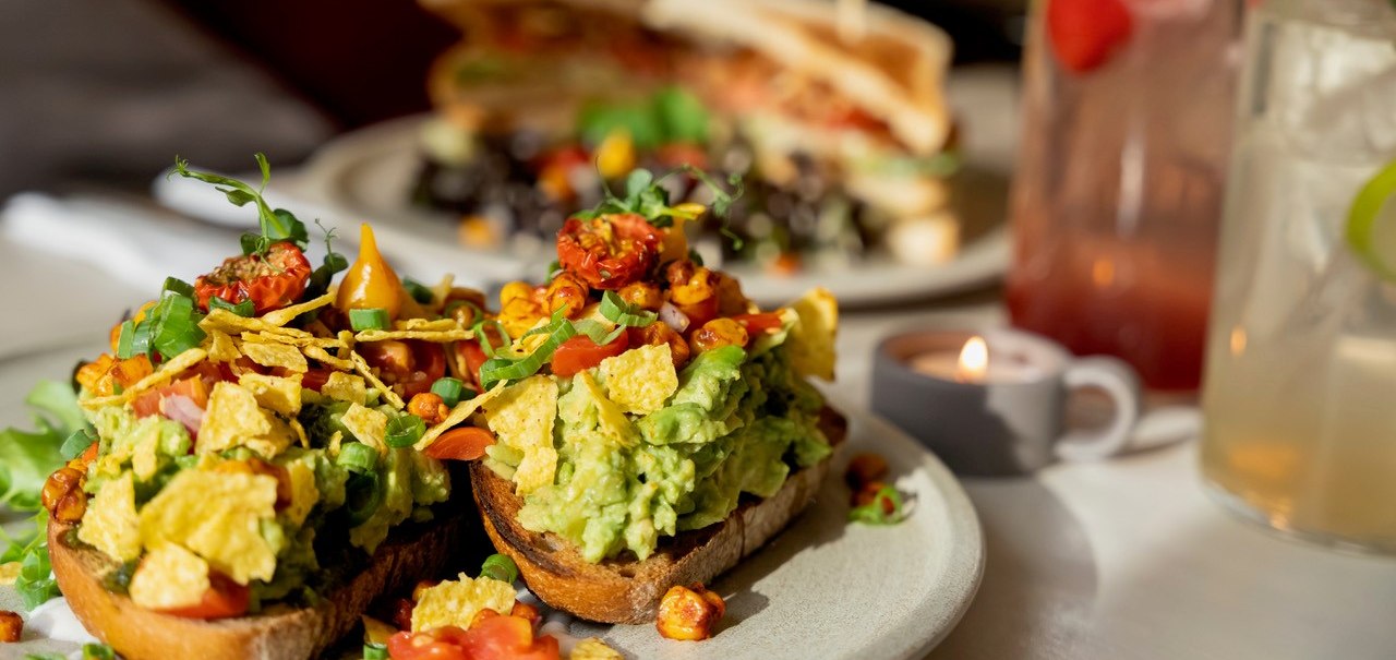Avocado toast with tomatoes and herbs, a sandwich and drinks in the background. A tea light creates a cozy atmosphere., © Veganes Gold®