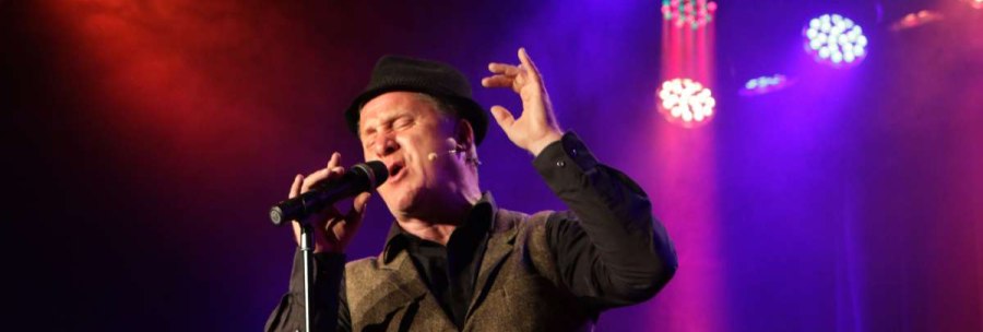 A singer in a hat performs passionately on a stage surrounded by colorful lights., © Sabine Haymann A singer in a hat performs passionately on a stage surrounded by colorful lights., © Sabine Haymann