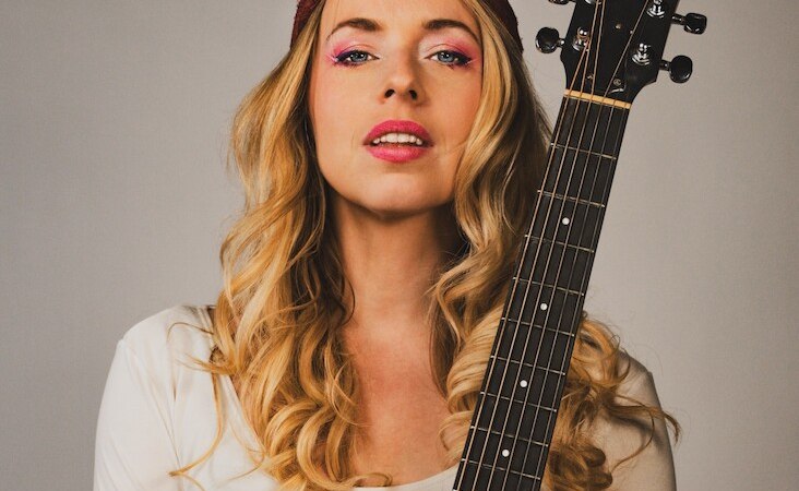 Frau mit blonden Locken, rotem Stirnband und Gitarre vor neutralem Hintergrund. Sie trägt ein weißes Oberteil und hat pinken Lidschatten., © Rosenau Kultur e.V. Frau mit blonden Locken, rotem Stirnband und Gitarre vor neutralem Hintergrund. Sie trägt ein weißes Oberteil und hat pinken Lidschatten., © Rosenau Kultur e.V.