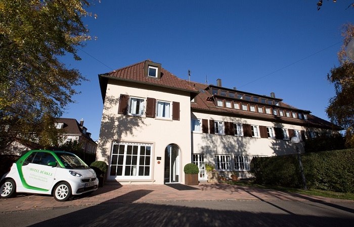 Ein zweistöckiges Hotel mit braunen Fensterläden, umgeben von Bäumen. Ein kleines Auto steht davor. Der Himmel ist klar und blau., © Hotel Bürkle