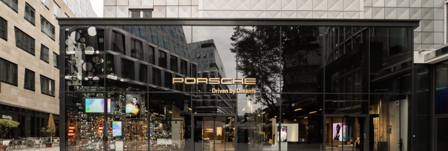 Porsche Brand Store with large glass façade, surrounded by modern buildings. The "Porsche Driven by Dreams" lettering is prominently visible., © Porsche AG