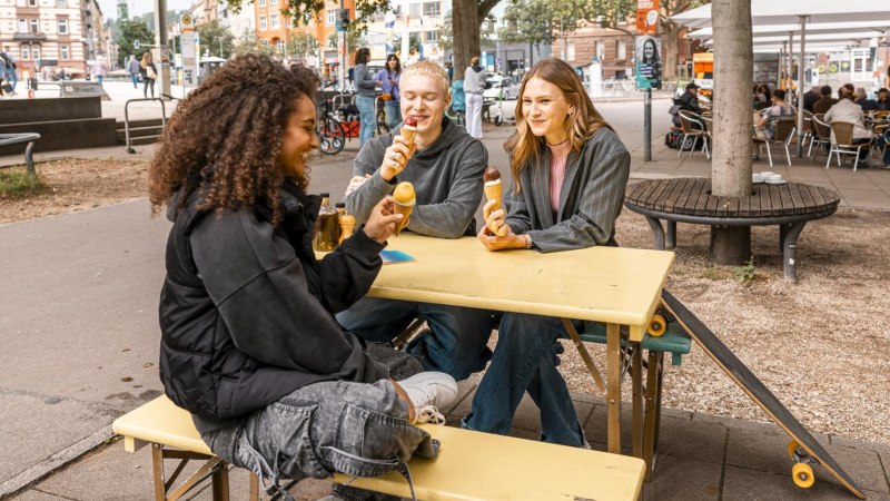 Drei Personen sitzen an einem gelben Tisch im Freien und essen Eis. Im Hintergrund sind Bäume, Fahrräder und ein Café zu sehen., © Stuttgart Marketing GmbH, Sarah Schmid