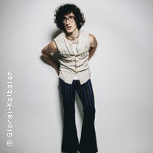 Person mit lockigem Haar und Brille, trägt ärmelloses Hemd und gestreifte Schlaghose, steht vor weißem Hintergrund., © links im Bild