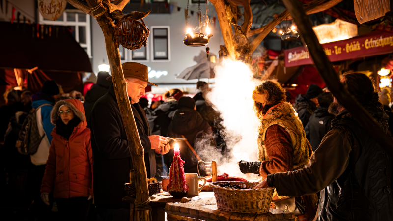 Belebter Weihnachtsmarkt mit Menschen, die an einem Stand mit Kerzen und dampfendem Essen stehen. Festliche Beleuchtung und geschäftige Atmosphäre., © Stuttgart-Marketing GmbH, Sarah Schmid