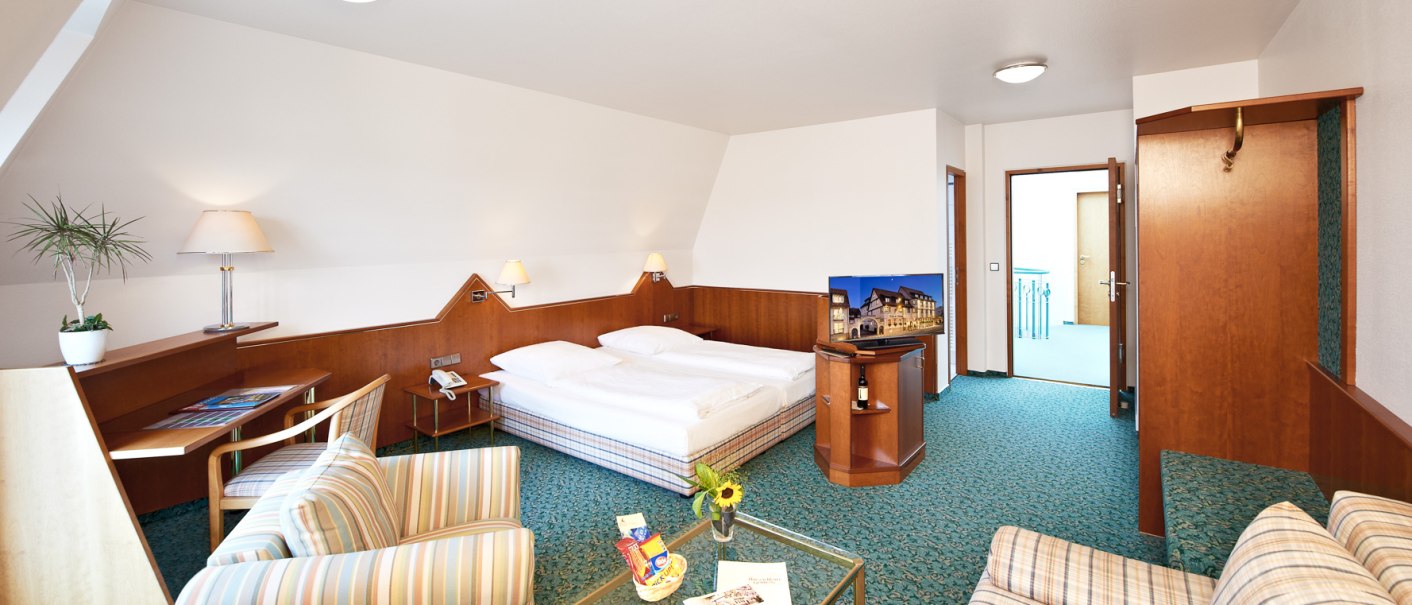 Geräumige Junior Suite mit Doppelbett, Sitzecke, Schreibtisch und Fernseher. Helle Einrichtung mit Holzmöbeln und grüner Teppichboden., © Hirsch Hotel