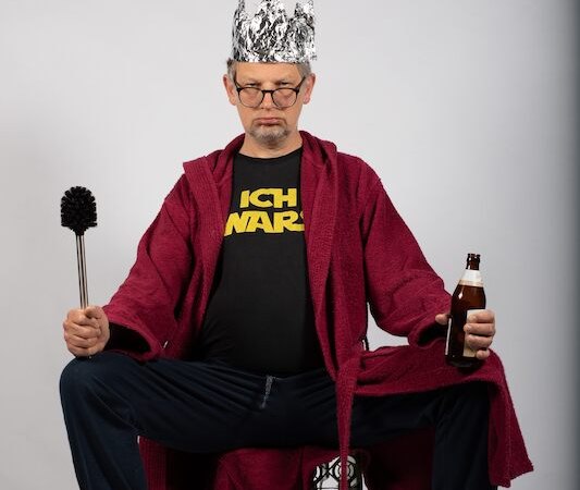 Person in rotem Bademantel und Alufolienkrone, hält eine Toilettenbürste und eine Bierflasche. T-Shirt mit Aufschrift 'ICH WARS'. Hintergrund neutral., © Rosenau Kultur e.V. Person in rotem Bademantel und Alufolienkrone, hält eine Toilettenbürste und eine Bierflasche. T-Shirt mit Aufschrift 'ICH WARS'. Hintergrund neutral., © Rosenau Kultur e.V.