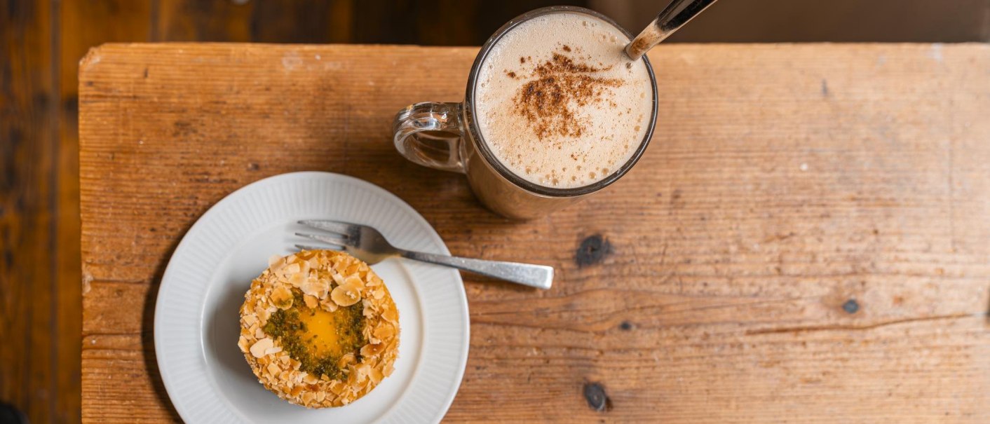 Ein Teller mit einem Geb&auml;ck, garniert mit Mandeln, und eine Tasse Kaffee mit Milchschaum auf einem rustikalen Holztisch., &copy; SMG, Sarah Schmid