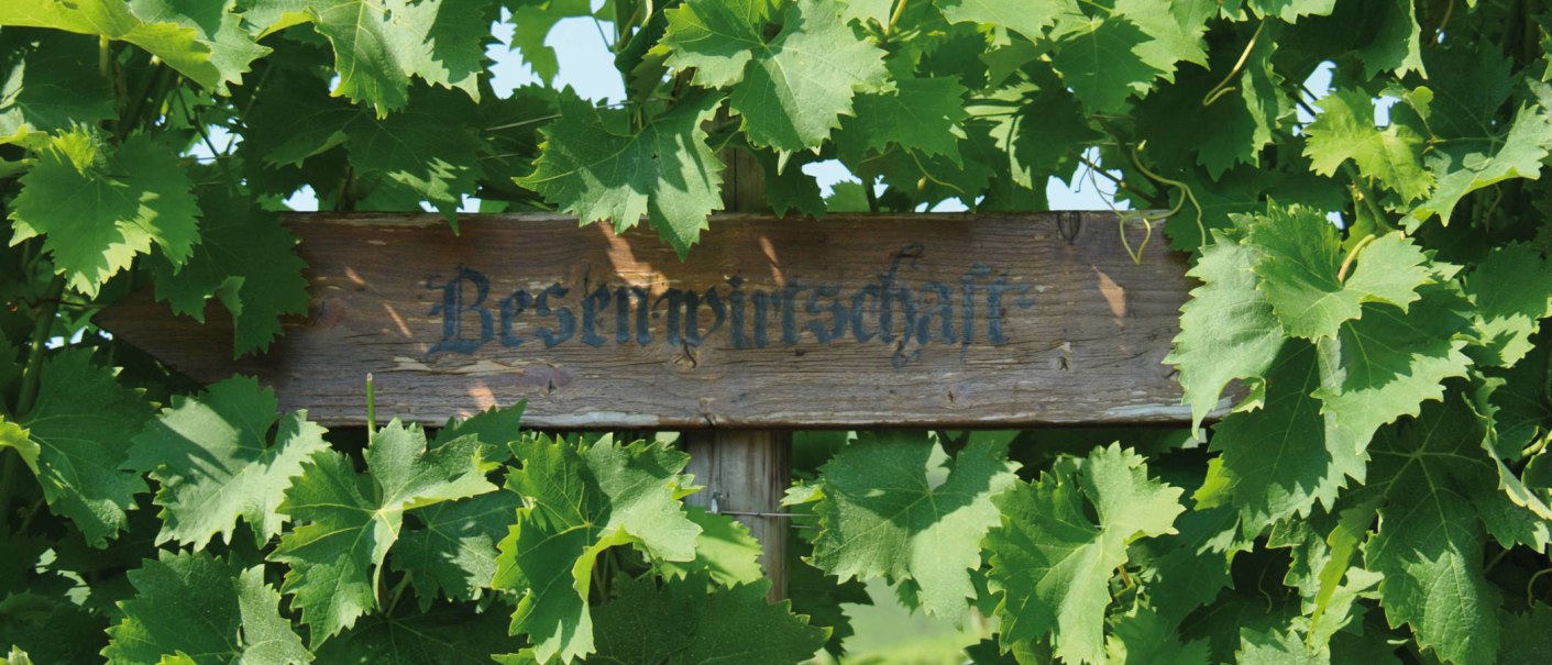 A wooden sign with the inscription 'Besenwirtschaft' is surrounded by green vine leaves., © SMG