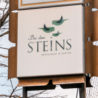A sign for the restaurant 'Bei den Steins' with birds as a logo, in front of a bare tree and blue sky., &copy; Stuttgart-Marketing GmbH, Sarah Schmid