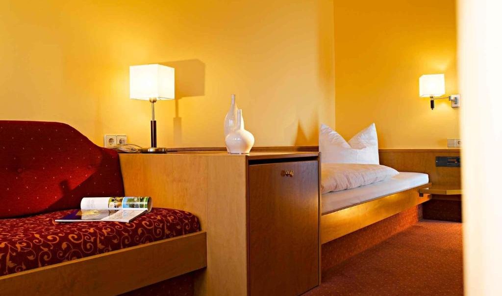A cozy hotel room with a single bed, red sofa, yellow walls and two lamps. A table with a vase and magazine can also be seen., © Nödinger Gastro GmbH