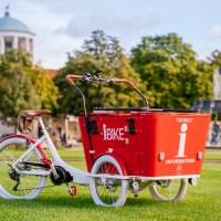 A red cargo bike with the inscription 'I-Bike' is parked on a meadow in front of a fountain and a building with a cupola., &copy; Stuttgart-Marketing GmbH, Thomas Niederm&uuml;ller