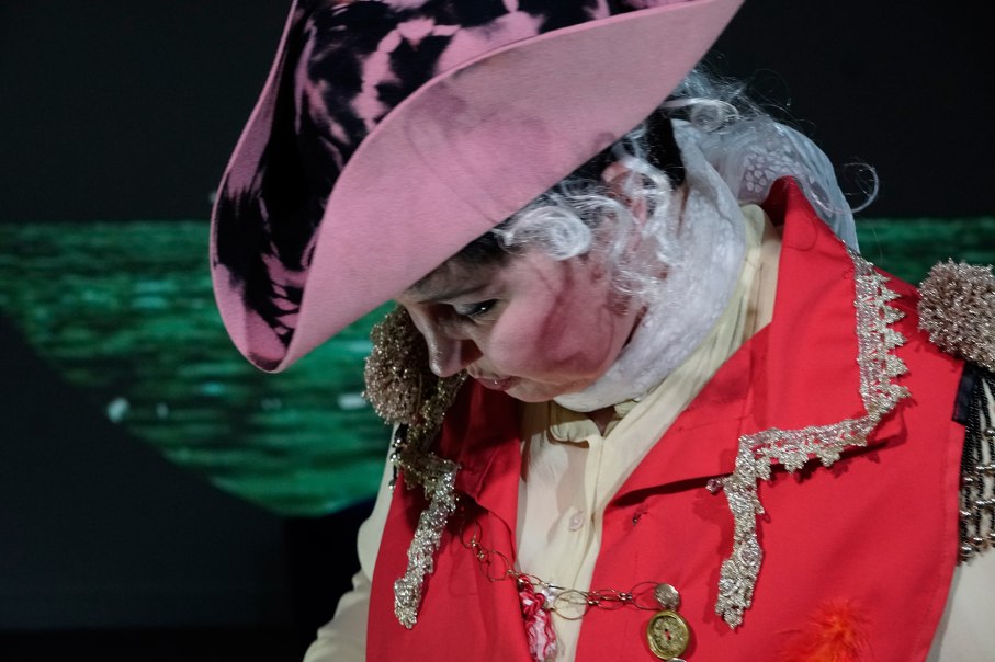 Eine Person in einem auff&auml;lligen historischen Kost&uuml;m mit rotem Mantel und rosa Hut, die nach unten schaut. Der Hintergrund ist dunkel und unscharf., &copy; Katja Ritter