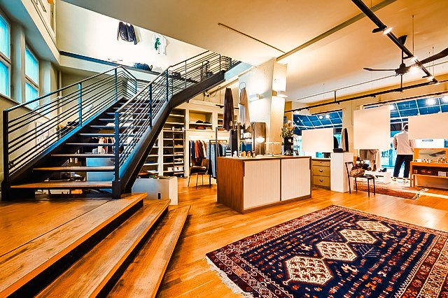Modernes Bekleidungsgeschäft mit offener Treppe, Holzboden und orientalischem Teppich. Kleidung und Accessoires sind stilvoll präsentiert., © Mario P. Rodrigues