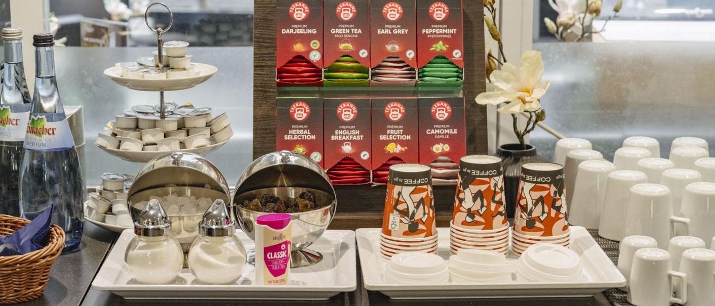 A breakfast buffet with various teas, bottles of water, sugar, cream and coffee mugs arranged on a table., © Plaza INN Stuttgart-Filderstadt A breakfast buffet with various teas, bottles of water, sugar, cream and coffee mugs arranged on a table., © Plaza INN Stuttgart-Filderstadt