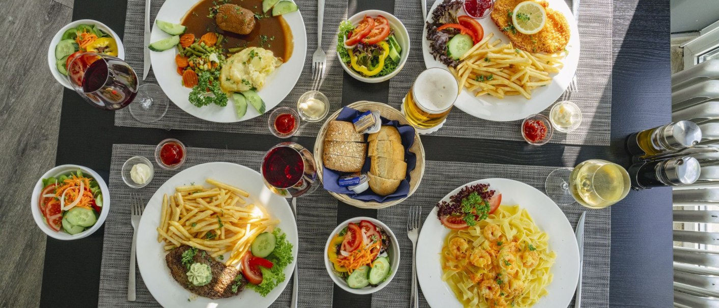 A table with various dishes: Meat, chips, salad, pasta, bread and drinks such as wine and beer in a restaurant., © Plaza INN Stuttgart-Filderstadt A table with various dishes: Meat, chips, salad, pasta, bread and drinks such as wine and beer in a restaurant., © Plaza INN Stuttgart-Filderstadt