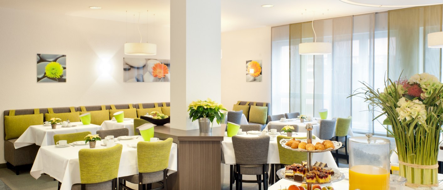 A bright breakfast room with set tables, green chairs, and a buffet featuring juice, pastries, and fruit. Flowers and pictures decorate the room., &copy; TOMAS