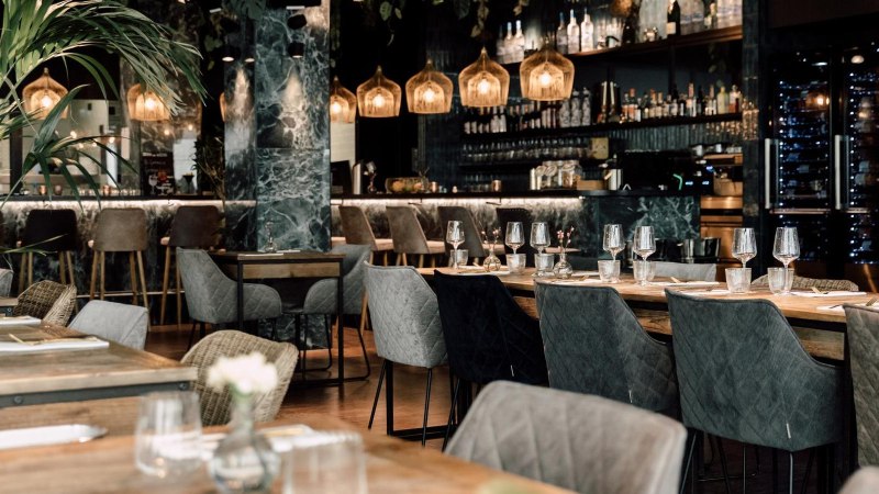 Elegant restaurant with modern furniture, laid tables and a stylish bar in the background. Subtle lighting and plants create a cozy atmosphere., © Malo Stuttgart - Lea Prestle