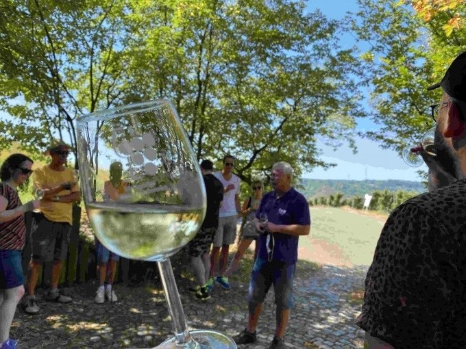 Gruppe bei einer Weinverkostung im Freien. Ein Glas Wei&szlig;wein wird in den Vordergrund gehalten. B&auml;ume und blauer Himmel im Hintergrund., &copy; Weingenuss Stuttgart