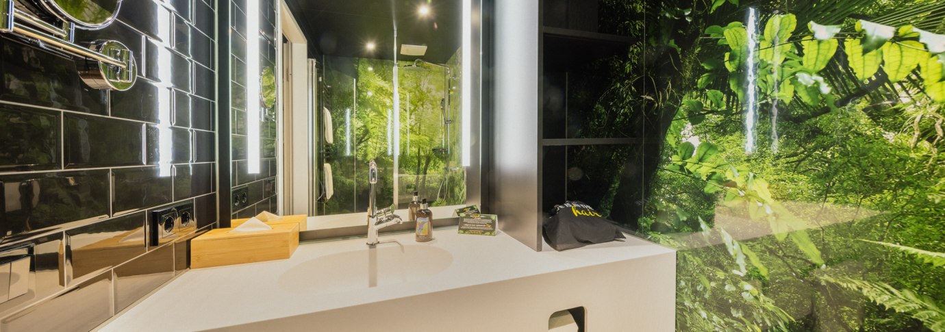 Modernes Badezimmer mit wei&szlig;em Waschbecken, Spiegel und Dschungelmotiv an der Wand. Schwarze Fliesen und helle Beleuchtung erg&auml;nzen das Design., &copy; Rioca Stuttgart Posto 4 GmbH