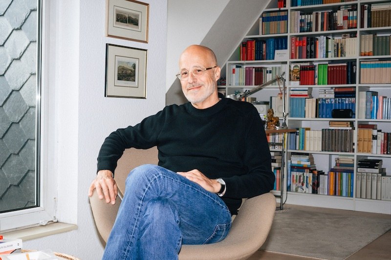 A man sits relaxed in a modern armchair in front of a large bookshelf. The room looks cozy and inviting., &copy; Theaterhaus Stuttgart e.V.