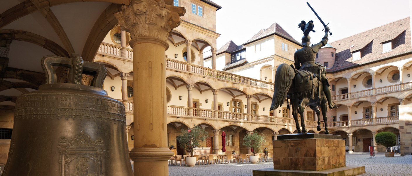 Inner courtyard of an old castle with equestrian statue, large bell and arcades. Tables and chairs stand in the courtyard, surrounded by historic buildings., © Stuttgart-Marketing GmbH, Christoph Düpper Inner courtyard of an old castle with equestrian statue, large bell and arcades. Tables and chairs stand in the courtyard, surrounded by historic buildings., © Stuttgart-Marketing GmbH, Christoph Düpper