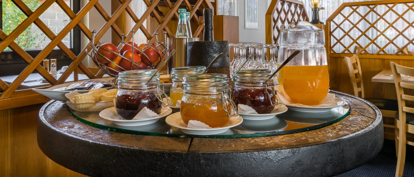 Ein Frühstückstisch mit Marmeladengläsern, einem Krug mit Orangensaft und einem Obstkorb voller Äpfel, umgeben von Holzmöbeln., © Novum Management GmbH Ein Frühstückstisch mit Marmeladengläsern, einem Krug mit Orangensaft und einem Obstkorb voller Äpfel, umgeben von Holzmöbeln., © Novum Management GmbH