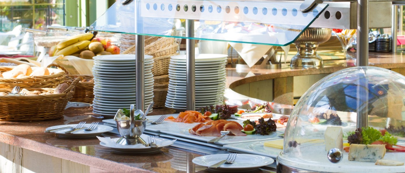 A rich buffet with plates, fruit, bread and cheese under a glass cover in a bright room with plants in the background., © TOMAS