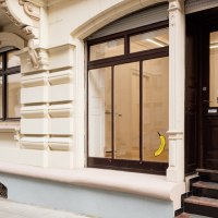 Eingang einer Galerie mit dem Schild 'Die Raumgalerie'. Eine Banane ist an der Glastür angebracht. Das Gebäude hat eine verzierte Fassade., © Dirk Wilhelmy Fotografie