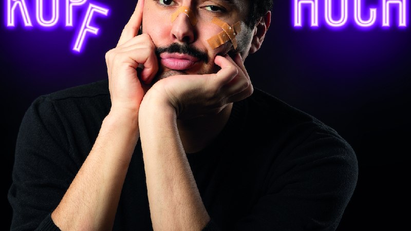 Ein Mann mit Pflastern im Gesicht stützt seinen Kopf in die Hände. Der Schriftzug 'Kopf hoch!' leuchtet in Neonfarben im Hintergrund., © SBEntertainment GmbH & Co. KG