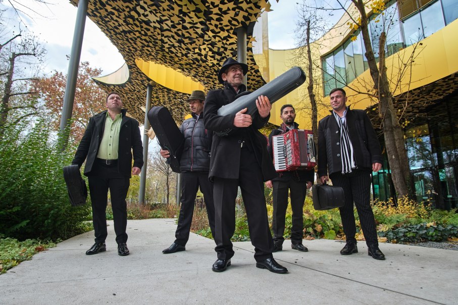 Fünf Musiker mit Instrumentenkoffern posieren vor einem modernen Gebäude mit gelbem, gemustertem Dach. Einer hält ein Akkordeon., © Szászcsávási Band Fünf Musiker mit Instrumentenkoffern posieren vor einem modernen Gebäude mit gelbem, gemustertem Dach. Einer hält ein Akkordeon., © Szászcsávási Band