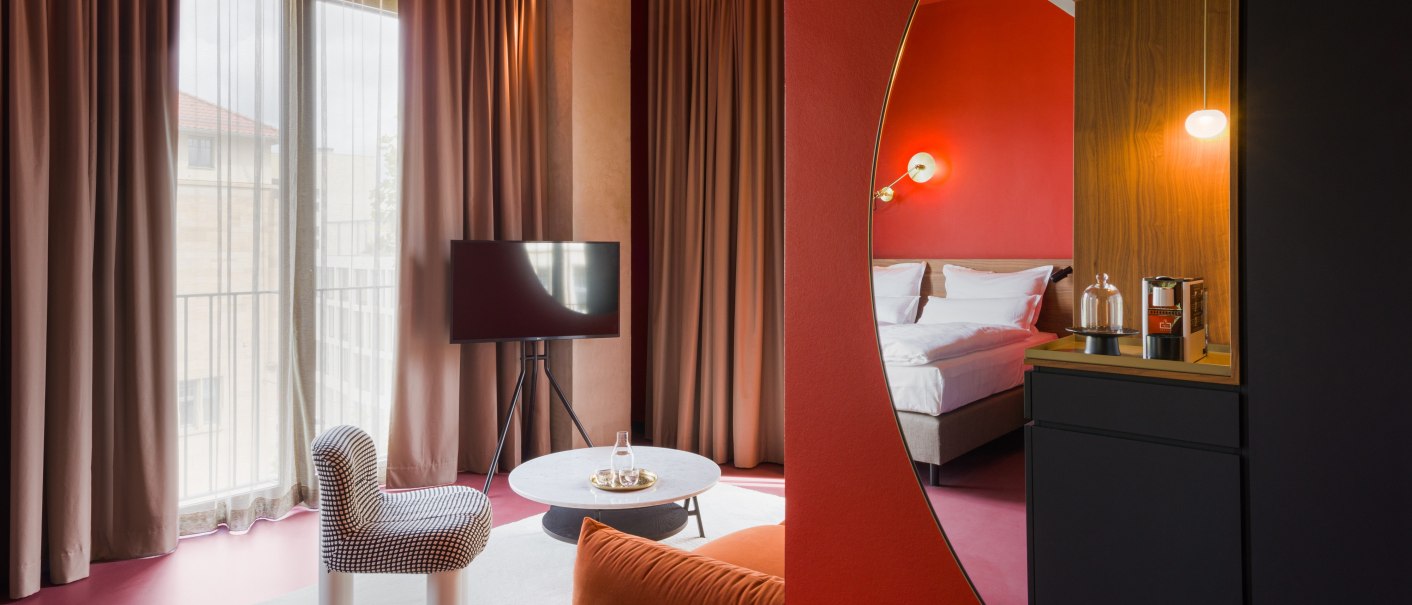 Moderne Hotelsuite mit roten Wänden, gemütlichem Bett, Sitzbereich mit Sessel und Tisch, Fernseher und stilvoller Beleuchtung., © Emilu Hotel GmbH
