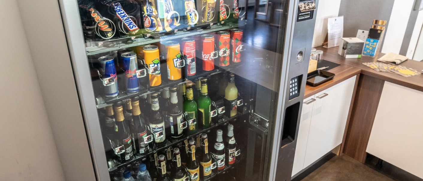 A vending machine in a hotel offers various drinks and snacks. A counter with information material can be seen in the background., &copy; a2 HOTELS