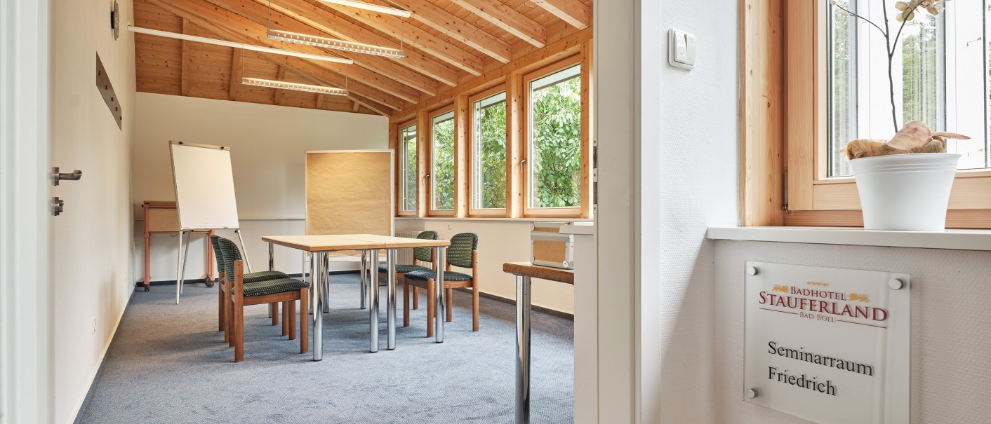 A bright seminar room with a wooden ceiling, a table, chairs, and a flip chart at the Badhotel Stauferland. A sign reading “Seminar Room Friedrich” is visible., © Badhotel Restaurant Stauferland A bright seminar room with a wooden ceiling, a table, chairs, and a flip chart at the Badhotel Stauferland. A sign reading “Seminar Room Friedrich” is visible., © Badhotel Restaurant Stauferland