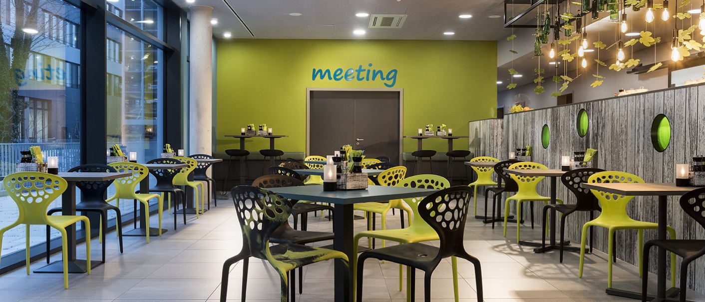 Modernes Restaurant mit grünen und schwarzen Stühlen, dekorativen Lichtern und Pflanzen. Große Fenster und eine grüne Wand mit 'meeting'-Schriftzug., © ibis Styles Stuttgart Vaihingen