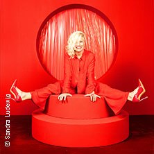 Woman in a red suit and red high heels sits smiling on a red pedestal in front of a red background., © links im Bild Woman in a red suit and red high heels sits smiling on a red pedestal in front of a red background., © links im Bild